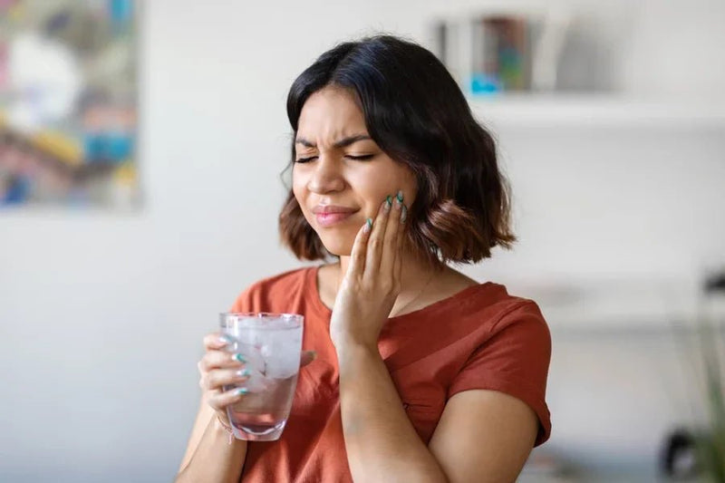 A young woman experiencing sharp pain from sensitive teeth while drinking a glass of iced water.