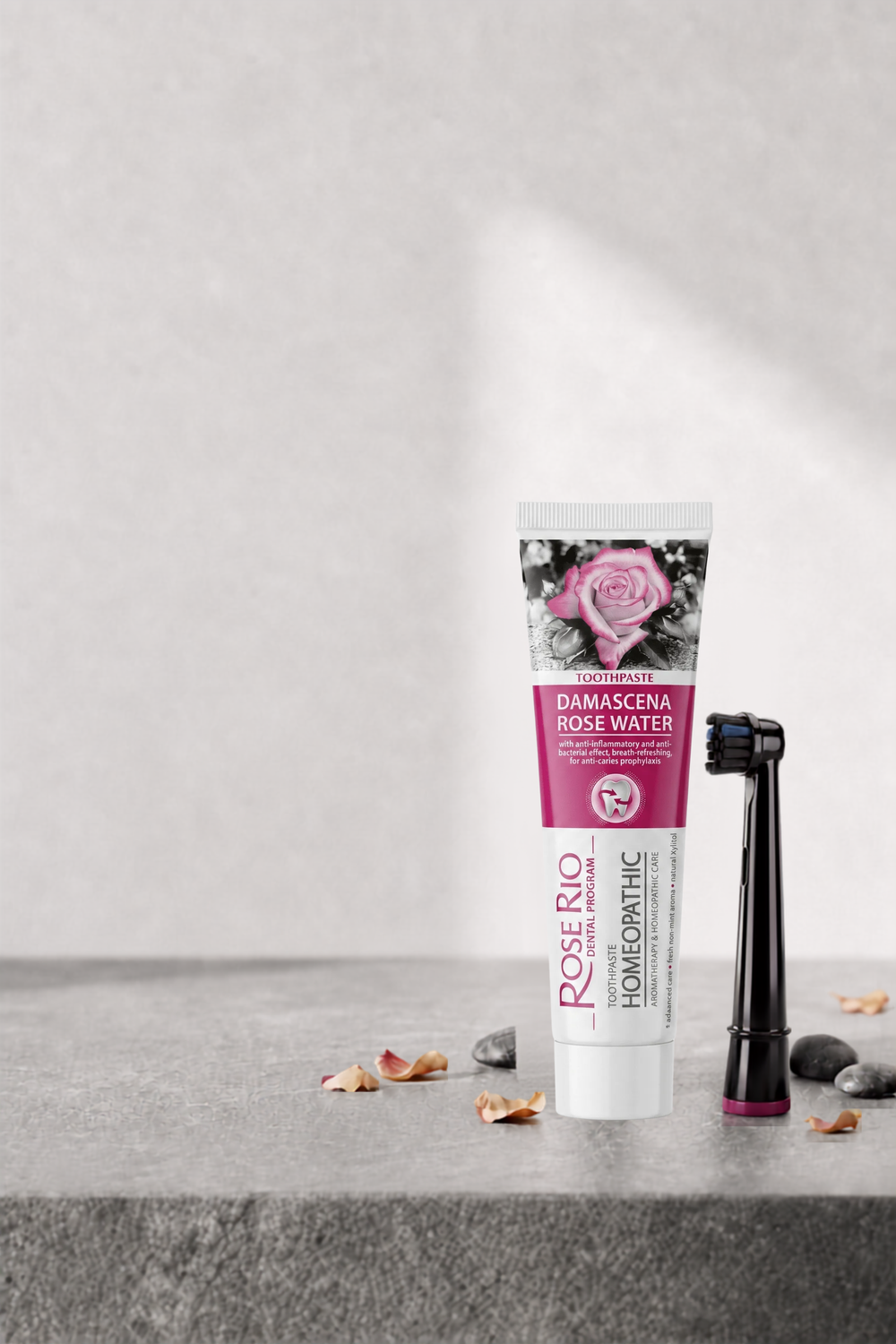 A tube of Rose Rio Home Spaface hair mask in Damascena Rose Water, displayed next to an electric toothbrush on a gray stone surface with decorative petals and stones.
