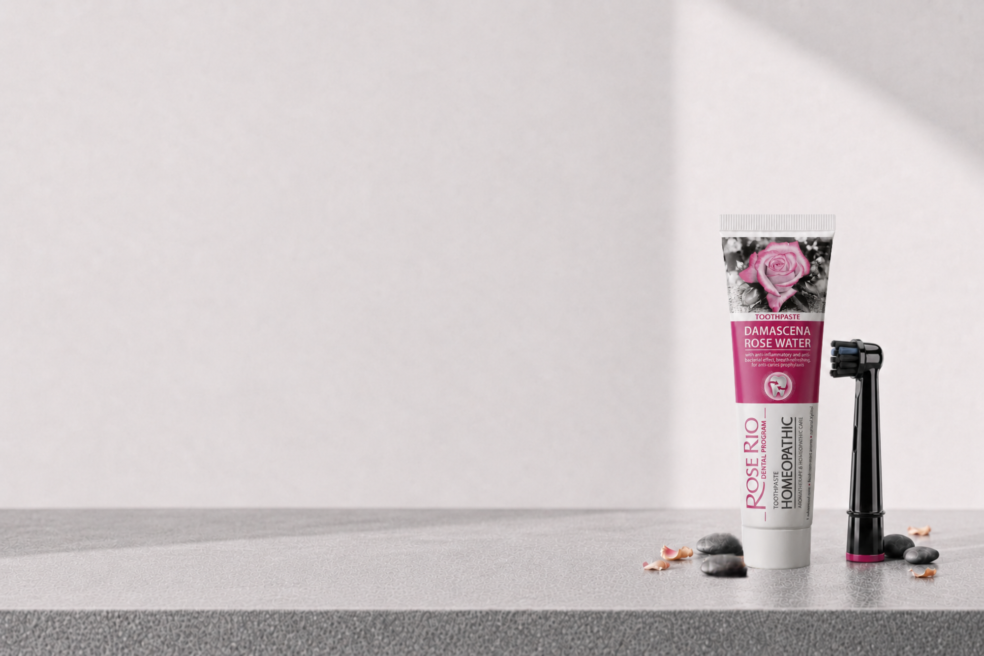 A tube of Rose Rio Dental Care toothpaste and a black electric toothbrush with a pink accent, styled on a gray stone surface against a light gray background.
